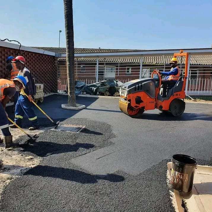 Ride-on roller compacting fresh asphalt in a parking area while the crew rakes the edges